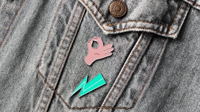 A denim jacket with two Project Me pins attached to the front pocket. One is a hand doing an okay sign. The other is a green lightening bolt.