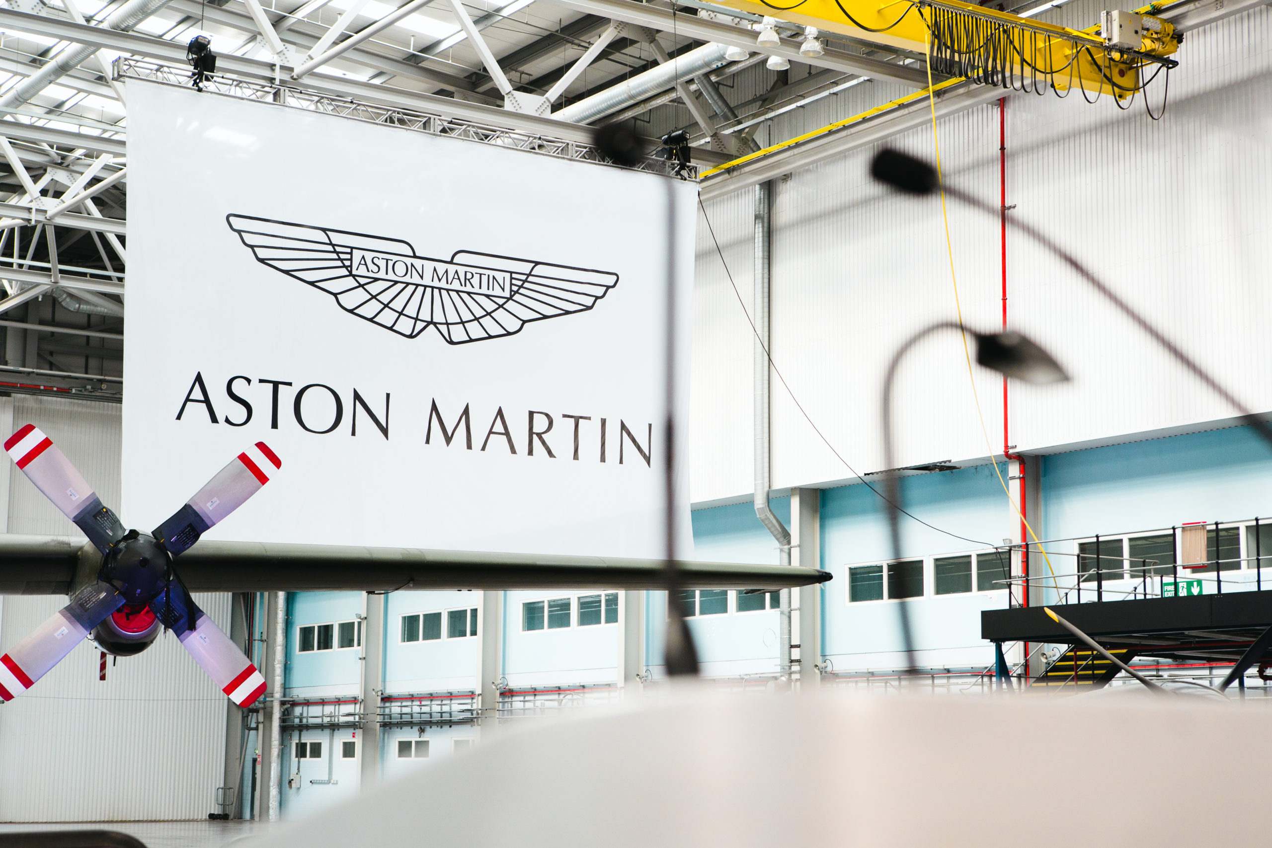 A view of a Aston Martin sign inside a hanger with a microphone out of focus and a wing of a plane in the background.