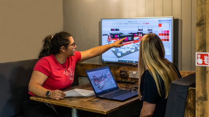 Two people having a meeting in a booth. One person is pointing at a screen with a 3d model showing.